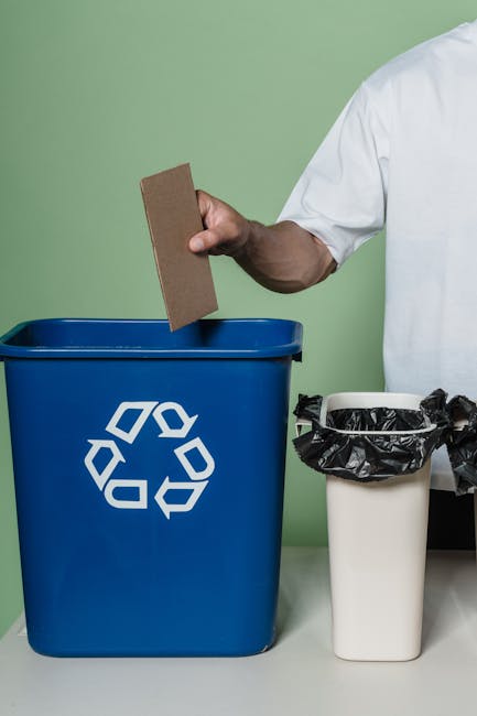 A green waste disposal bin with a black pictogram of a person discarding rubbish, mounted on metal supports adjacent to a yellow tiled wall exterior of a building. The bin has a rectangular open top a