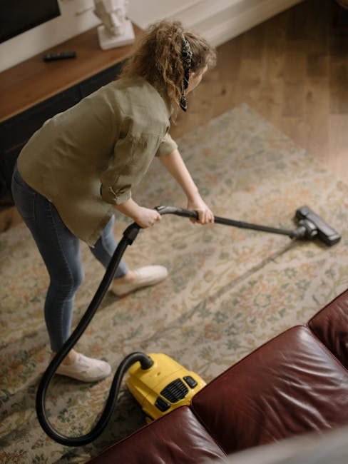 A person dressed in white protective coveralls, face mask, and blue disposable gloves is performing deep carpet cleaning in a bright living room. They are using a professional vacuum or steaming devic