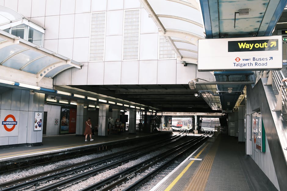 Exterior view of Hammersmith station in London, featuring railway tracks running through an underground area with a covered walkway on the side. The station has a modern design with white tiled walls,
