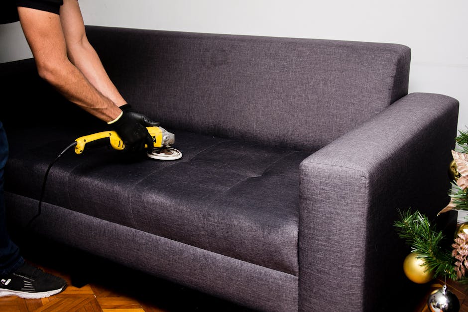 A person wearing black gloves is using a handheld electric rotary polisher on a dark grey fabric upholstered sofa, focusing on the seat cushion. The sofa is positioned in a bright room with white wall
